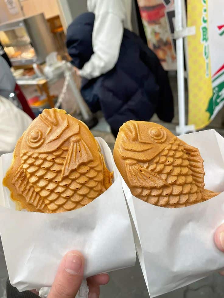 Two freshly made taiyaki pastries held in paper wrappers at a bustling market.