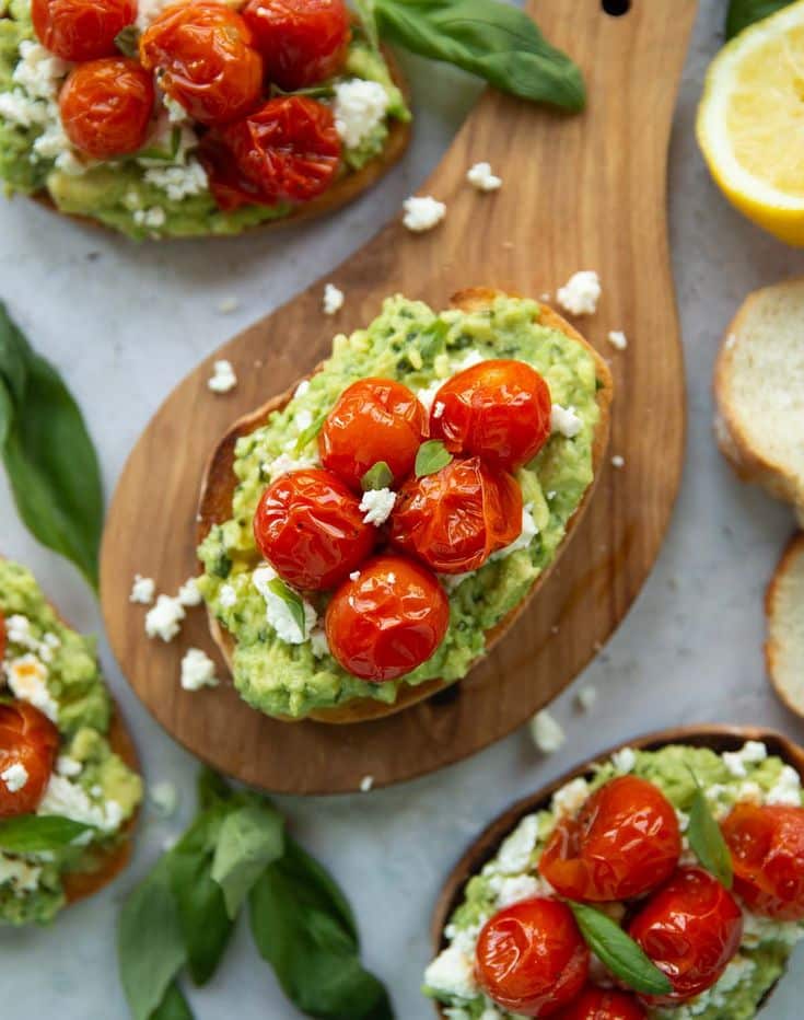 Avocado toast topped with roasted cherry tomatoes and feta on a wooden board, garnished with fresh basil leaves.