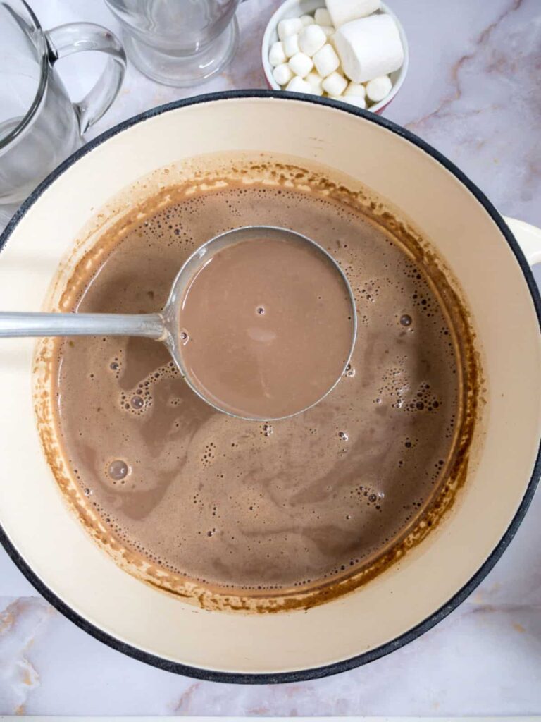 Creamy hot chocolate in a pot with a ladle, surrounded by clear mugs and marshmallows on a marble countertop.