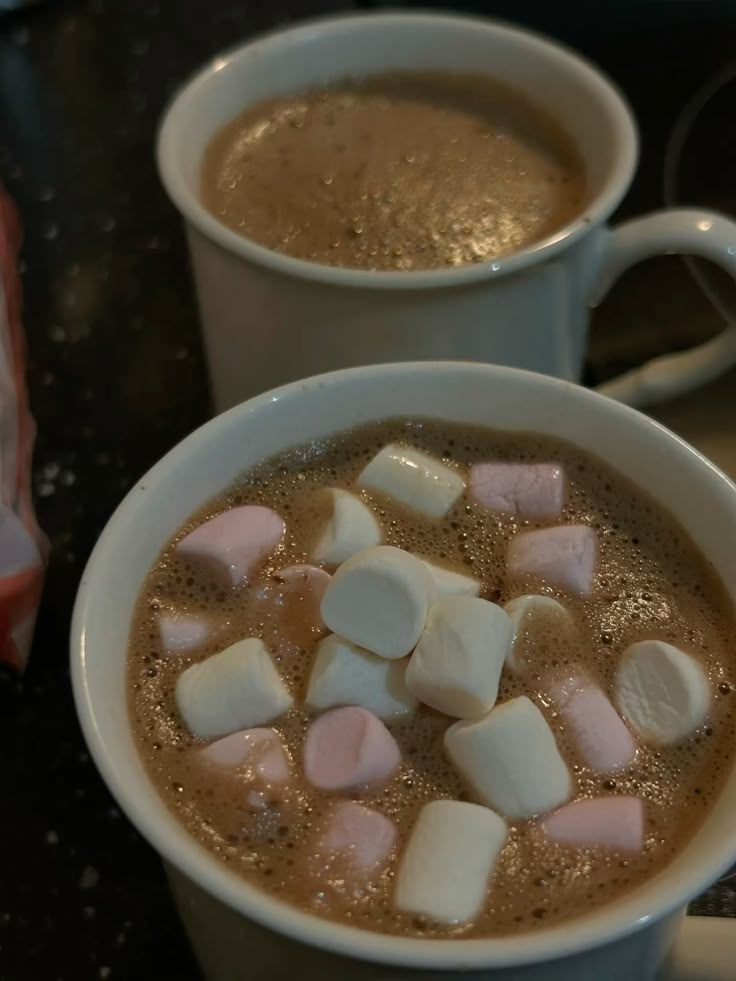 Two mugs of hot chocolate topped with colorful marshmallows, set on a dark countertop.