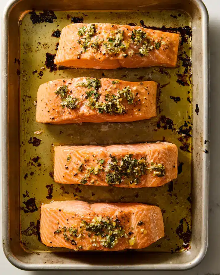 Four seasoned salmon fillets on a baking tray, ready to be cooked.