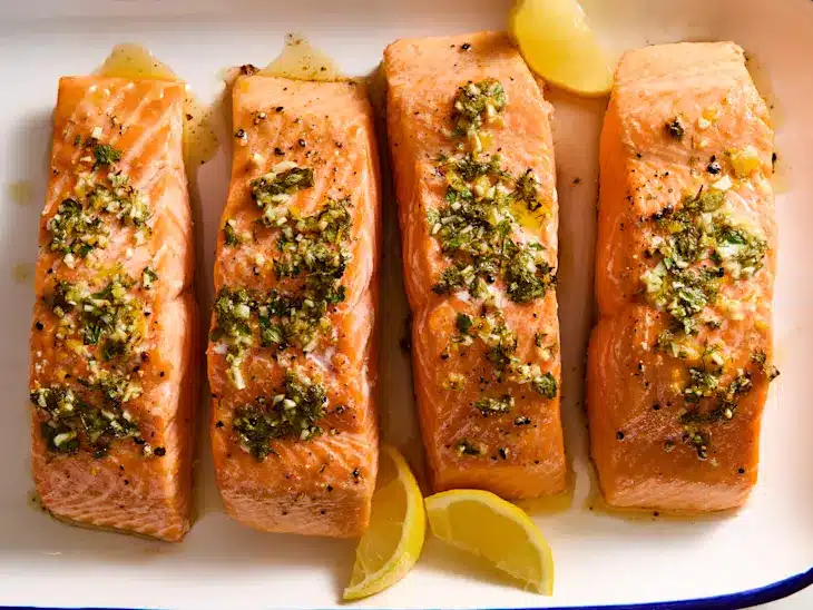 Four seasoned salmon fillets garnished with herbs, served on a plate with lemon wedges. Delicious seafood meal.