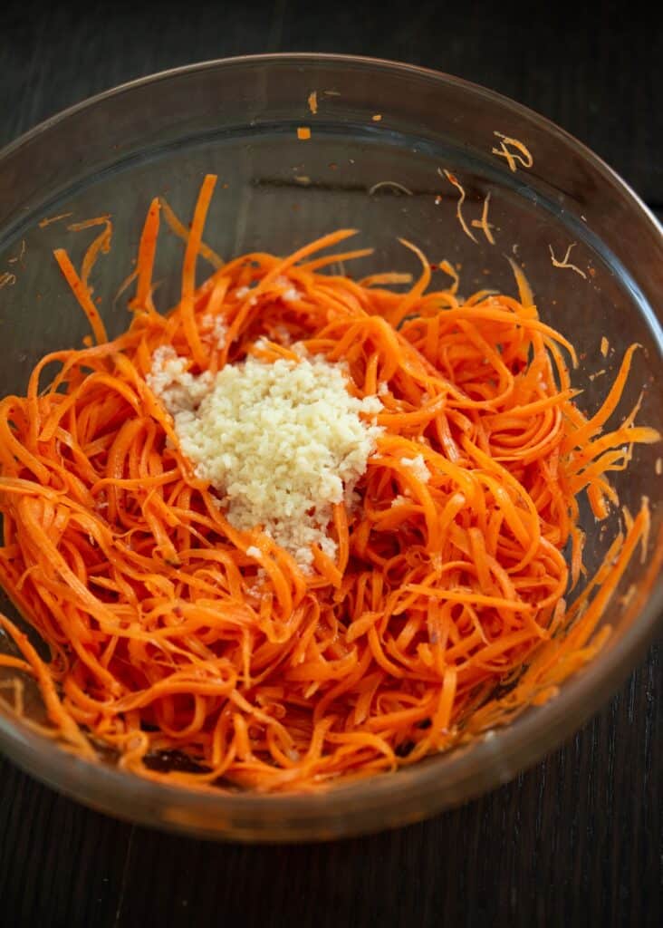 Shredded carrots with garlic in a glass bowl on a dark table, ready to be mixed for salad preparation.