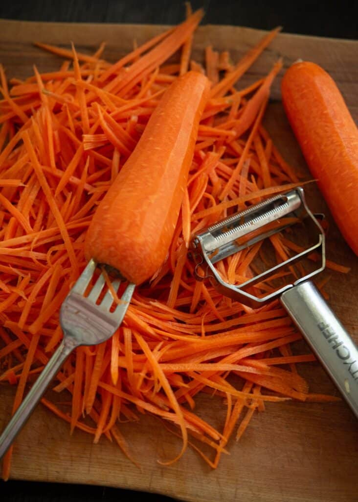 Freshly peeled carrots and shredded slices on a cutting board with a vegetable peeler and fork.