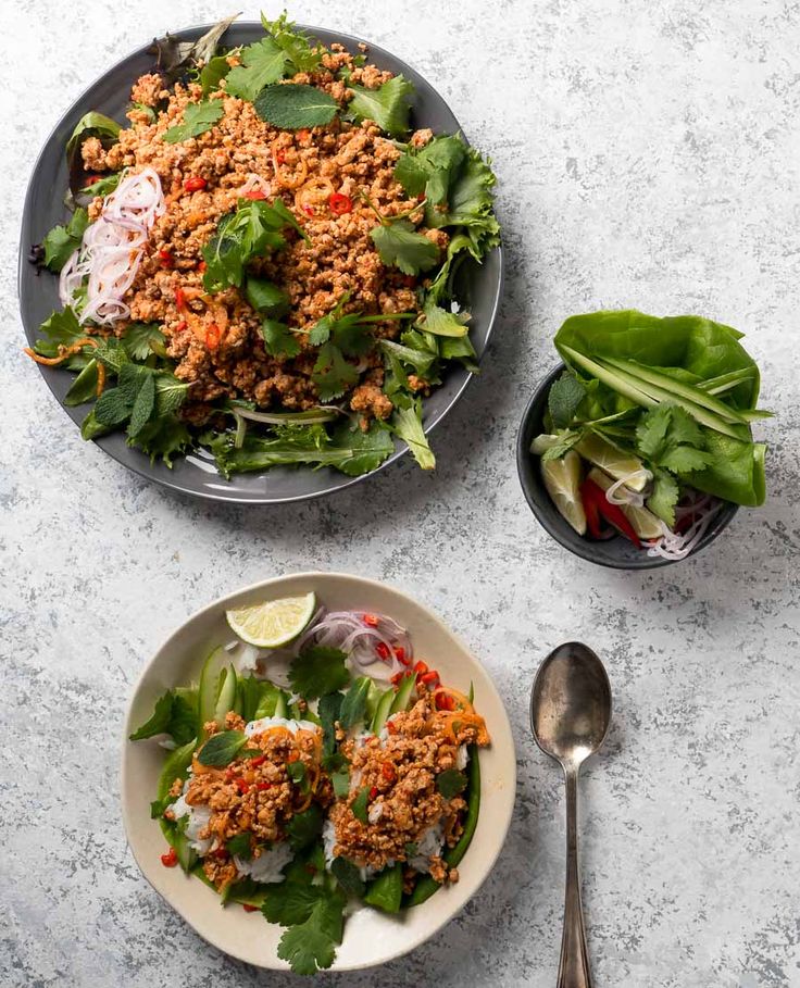 Delicious Thai larb salad with herbs, chilies, and lime on plates with fresh vegetable garnishes, served on a textured surface.