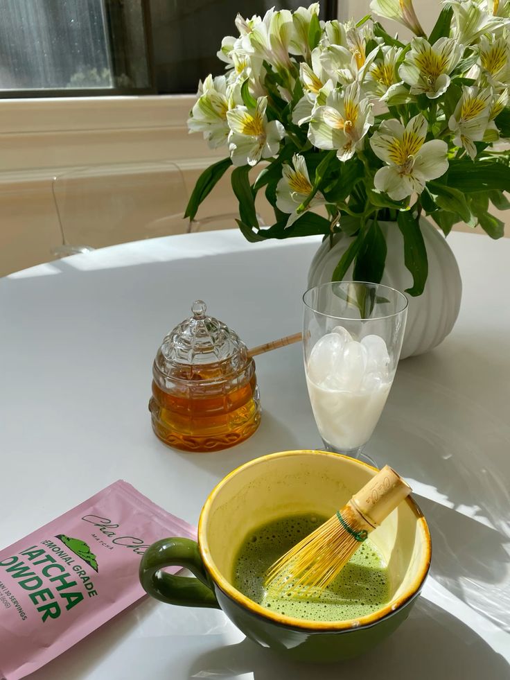 Green matcha tea in a mug with a whisk, honey jar, and flowers on a sunlit table.