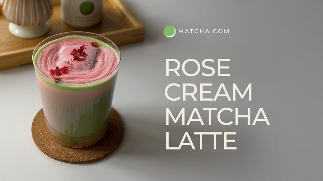 Rose cream matcha latte with pink foam and petals in a glass on a cork coaster, next to a wooden tray.