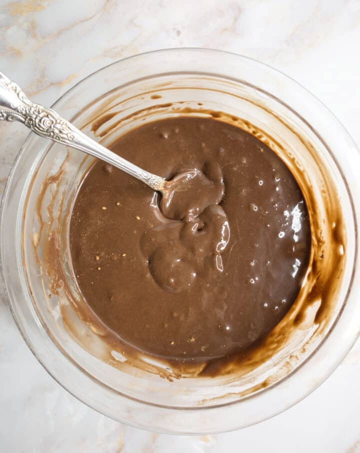 Chocolate cake batter in a glass bowl with a spoon on a marble countertop.