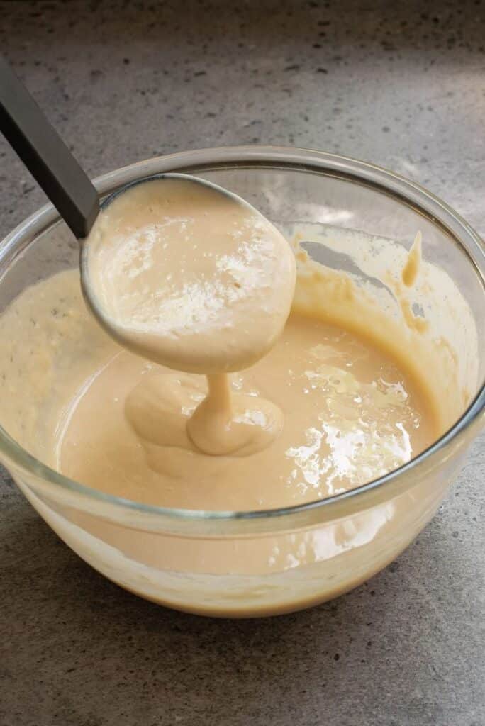 Thick pancake batter in a glass bowl, poured with a ladle, perfect for breakfast recipes.