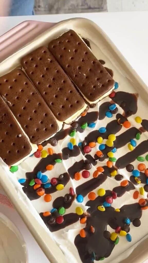 Ice cream sandwich cake topped with chocolate sauce and colorful candies in a baking dish.
