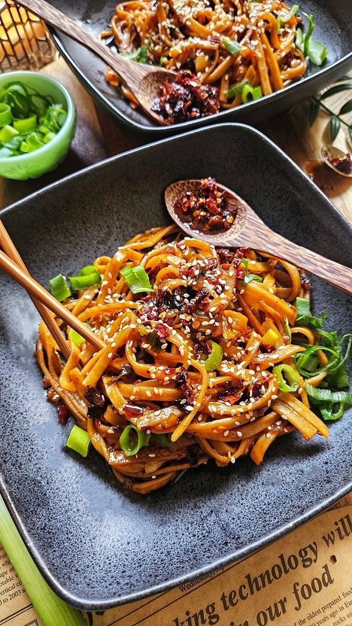 15 Minute Chili Garlic Noodles - Fiery Chili Garlic Noodles Recipe – Quick & Flavorful - Chief Stories Savory noodles with soy sauce, green onions, and sesame seeds in a black bowl, topped with spicy chili flakes.