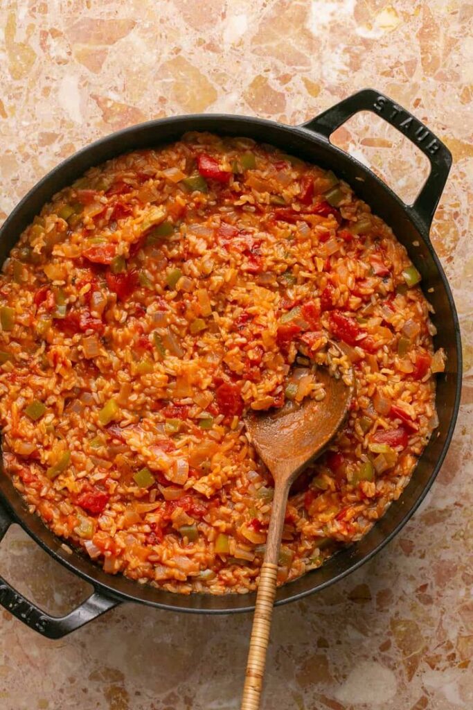 30-Minute Spanish Chicken and Rice One-Skillet - One-Pan Spanish Style Meat and Tomato Rice: Your New Weeknight Hero! | Spanish rice with tomatoes and green peppers in a black pot on a marble surface, stirred with a wooden spoon.