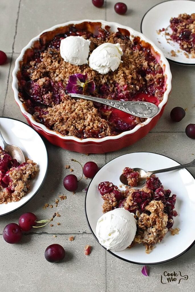 Cherry crumble with ice cream served in a dish and plates, topped with a flower, on a rustic table.