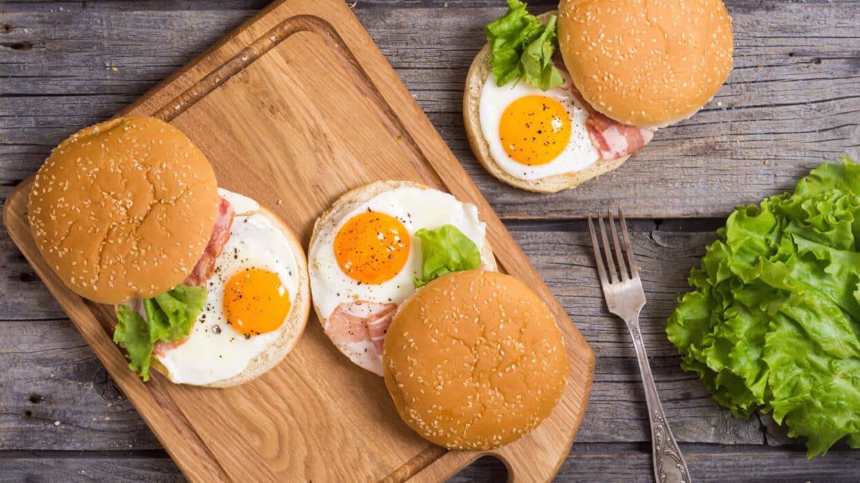 Three breakfast burgers with fried eggs, bacon, and lettuce on a wooden board, next to fresh lettuce and a fork.