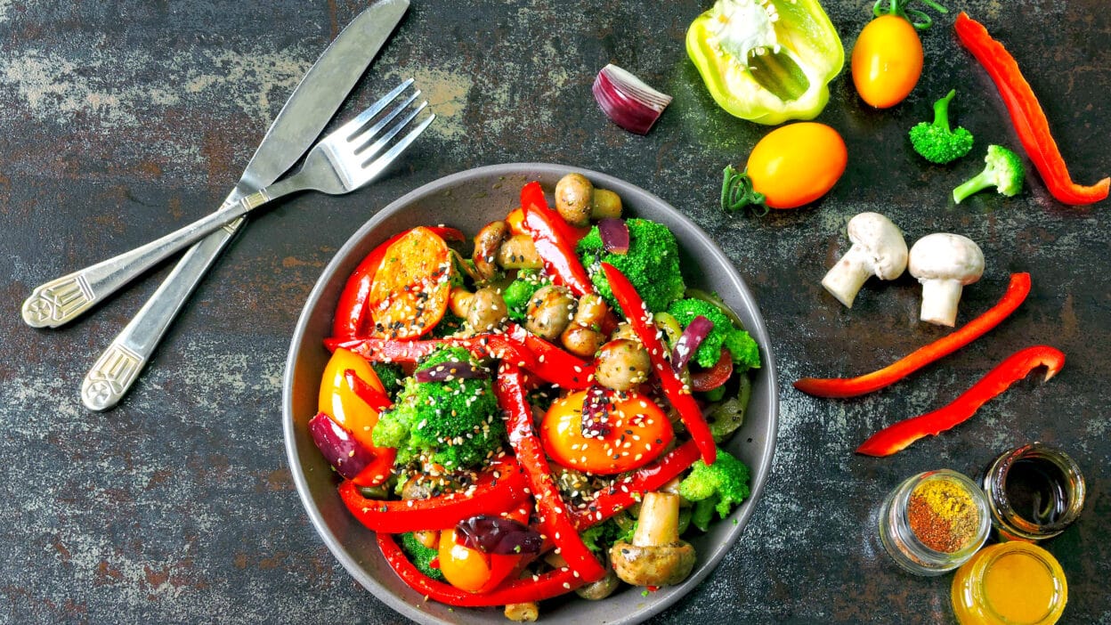 Colorful stir-fried vegetables in a bowl with spices and utensils on a rustic table.