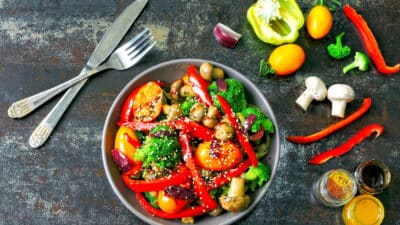 Colorful stir-fried vegetables in a bowl with spices and utensils on a rustic table.