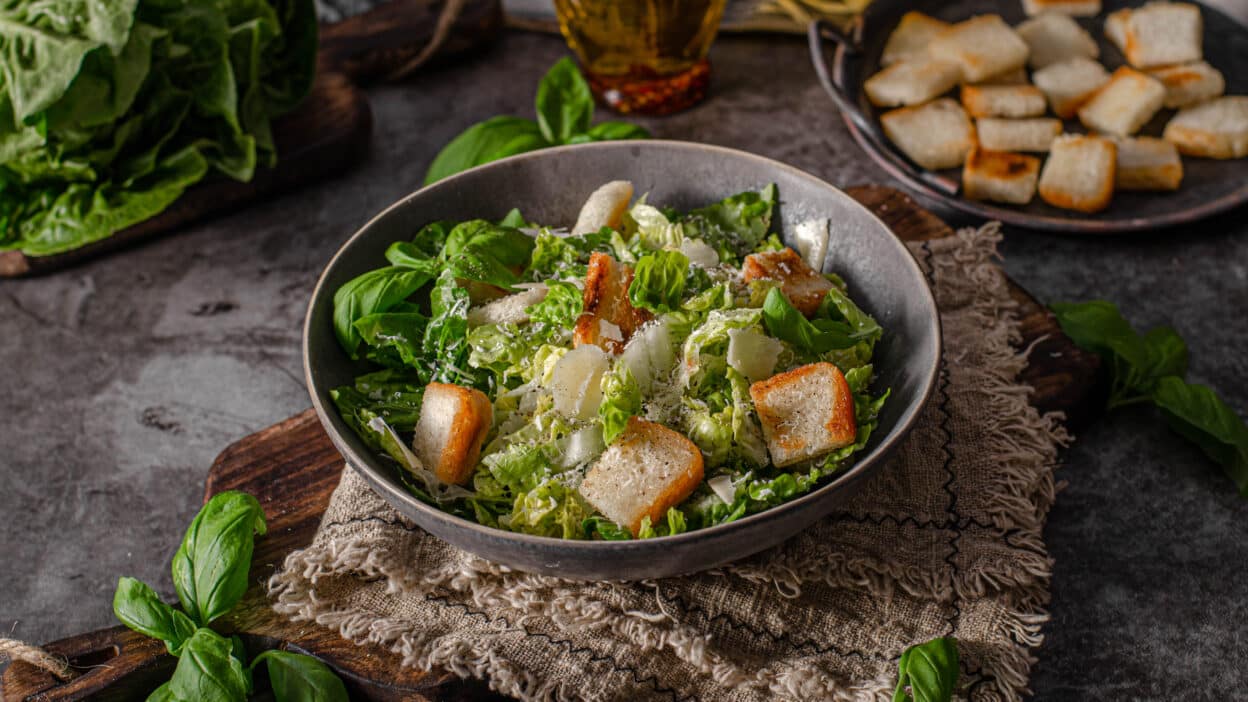 Fresh Caesar salad with croutons and parmesan in dark bowl on rustic table setting.