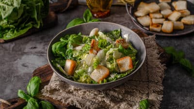 Fresh Caesar salad with croutons and parmesan in dark bowl on rustic table setting.