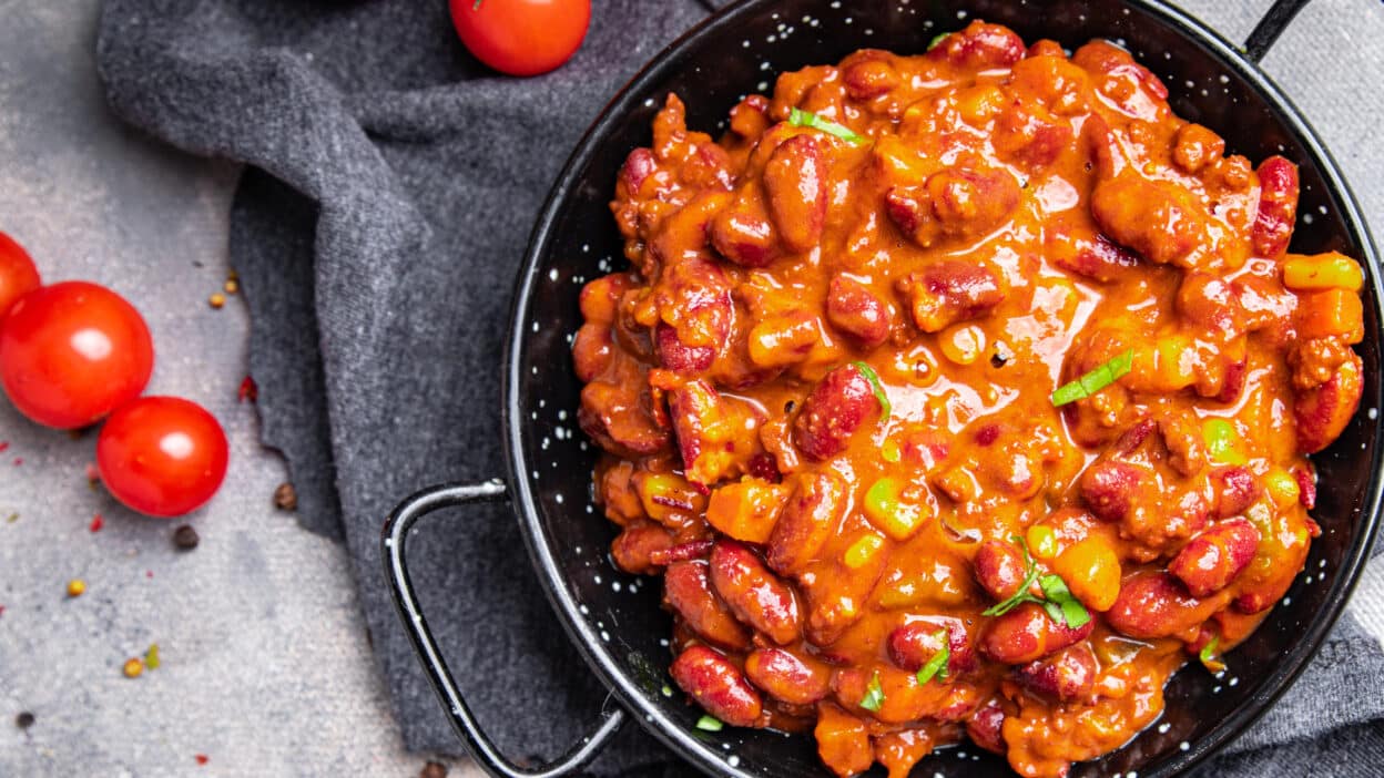 Hearty chili con carne in a black bowl garnished with fresh herbs, surrounded by cherry tomatoes and spices.
