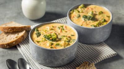 Creamy broccoli cheese soup in gray bowls with rustic bread on a striped cloth. Perfect for a cozy, comforting meal.