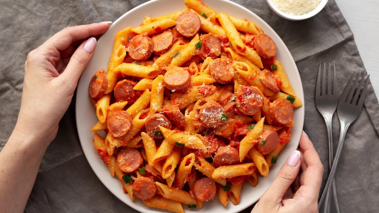 Plate of penne pasta with sausage and tomato sauce, garnished with cheese, held by hands next to forks on a napkin.