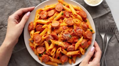 Plate of penne pasta with sausage and tomato sauce, garnished with cheese, held by hands next to forks on a napkin.
