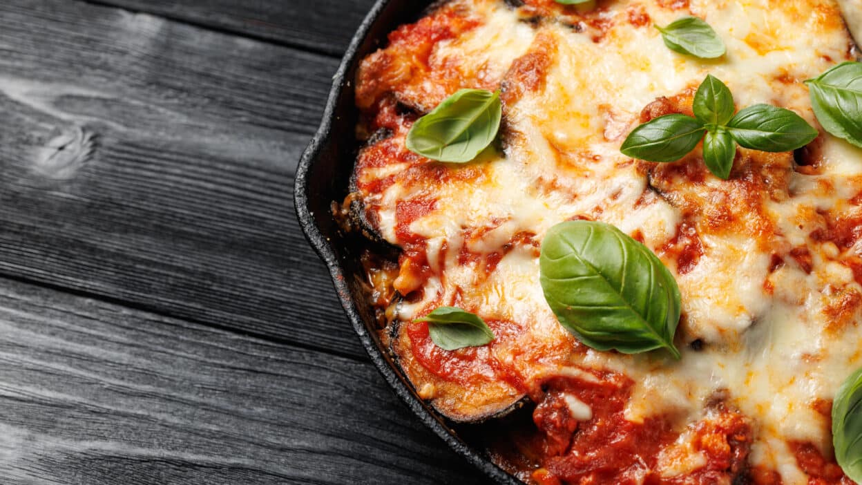 Cheesy eggplant Parmesan in skillet with fresh basil on rustic wooden table.