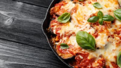 Cheesy eggplant Parmesan in skillet with fresh basil on rustic wooden table.