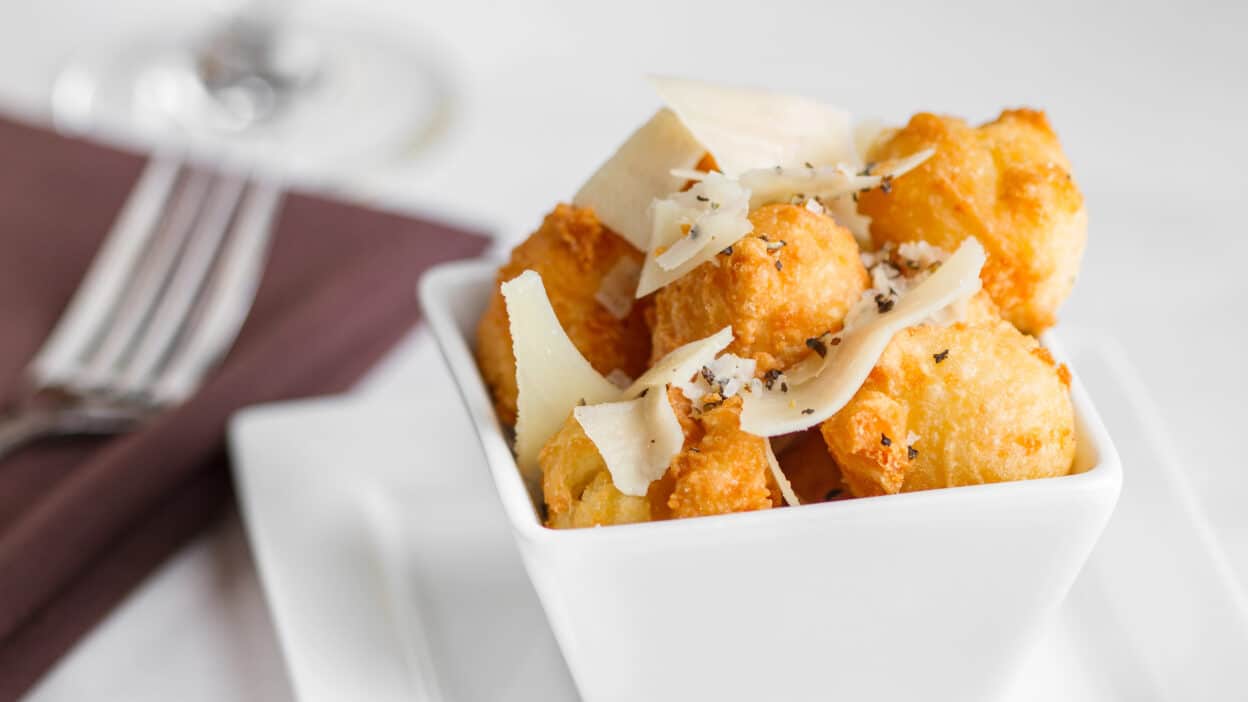 Crispy tater tots topped with shaved cheese in a white bowl, served with a fork and napkin on the side.