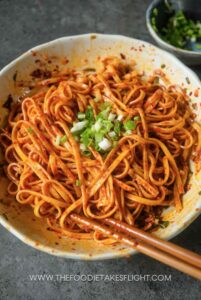 Easy Chili Garlic Oil Noodles - Fiery Chili Garlic Noodles Recipe – Quick & Flavorful - Chief Stories Spicy noodles topped with fresh green onions, served in a bowl with chopsticks on a gray surface.