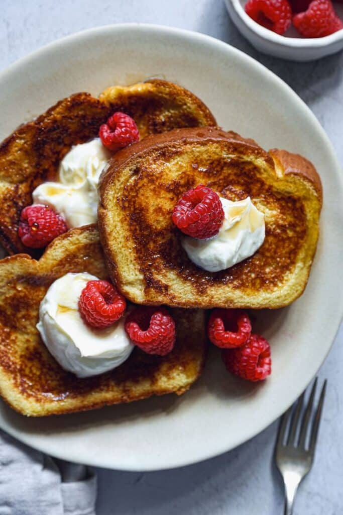French toast with fresh raspberries and cream on a beige plate, fork on the side. Delicious breakfast idea.