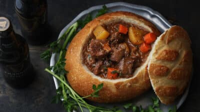 Hearty beef stew in a bread bowl with carrots and potatoes, garnished with parsley, next to bottles of stout beer.