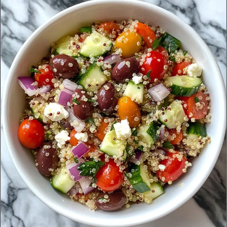 Mediterranean Quinoa Salad Bowl Recipe - Greek Island Flavors - Mediterranean Quinoa Salad Bowl: Fresh & Flavorful Quinoa salad with cherry tomatoes, cucumbers, olives, red onion, and feta in a white bowl on a marble surface.