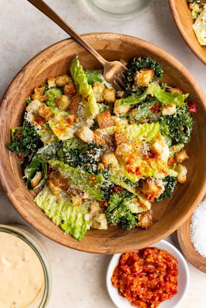 Spicy Caesar salad - The Bold Appetite - Classic Caesar Salad: Homemade Recipe - Chief Stories Fresh Caesar salad with croutons and Parmesan in a wooden bowl, accompanied by dipping sauces on the side.