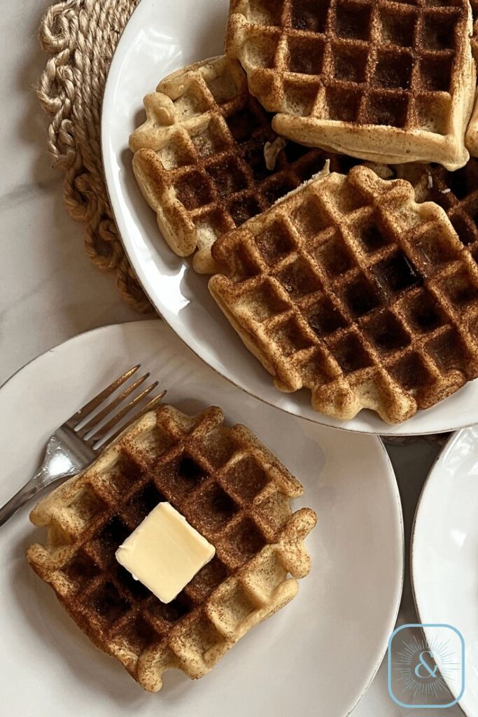 Golden brown waffles stacked on a plate, topped with a pat of butter, next to a fork on a white plate.
