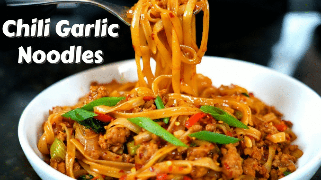 Spicy chili garlic noodles with vibrant vegetables and meat, served hot in a white bowl.