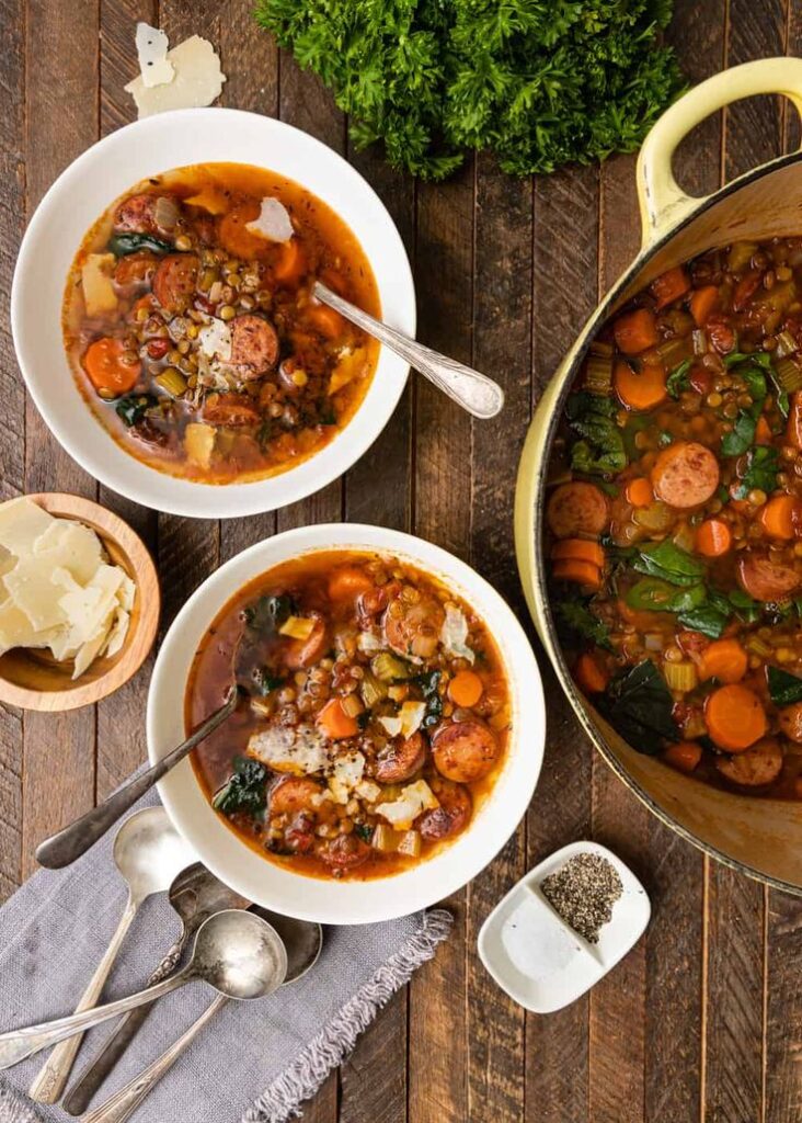 Bowls of hearty sausage and lentil soup on a wooden table, garnished with fresh herbs and parmesan.