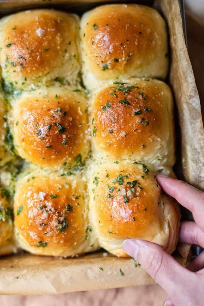 Bring Soft Buttery Garlic Parmesan Pull-Apart Rolls To the Celebration - Garlic Parmesan Pull-Apart Rolls Recipe Freshly baked garlic butter rolls with herbs in a baking tray, being picked up by hand.