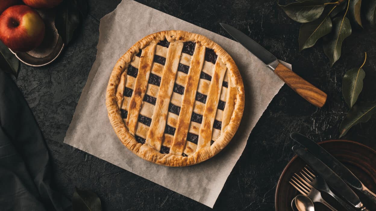 Depositphotos_166416512_XL - Classic Homemade Apple Pie Recipe Freshly baked lattice apple pie on parchment paper with apples and cutlery nearby.