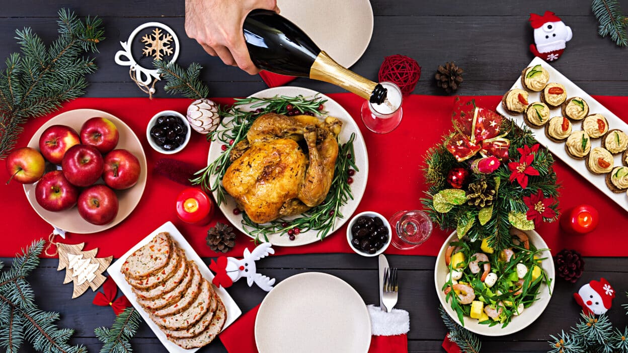 Festive holiday table with roasted chicken, apples, salad, and champagne. Christmas decorations and candles adorn the scene.