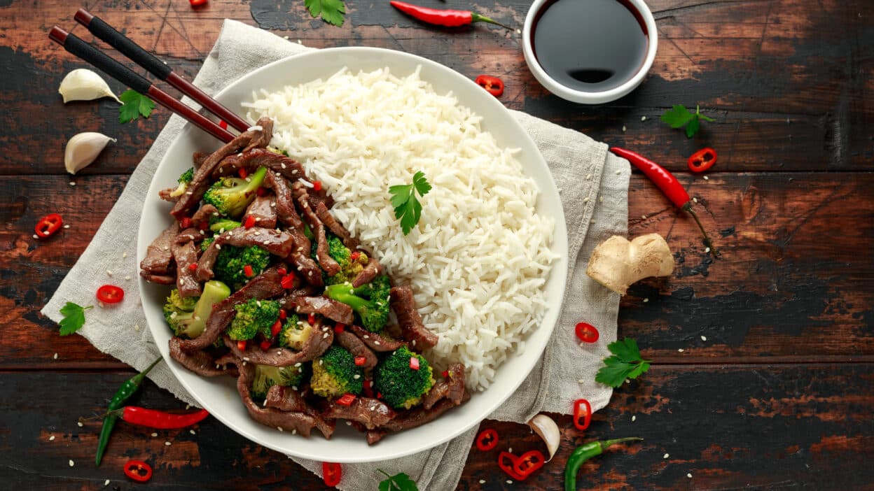 Plate of beef stir fry with broccoli and white rice on a rustic table with chopsticks and soy sauce.