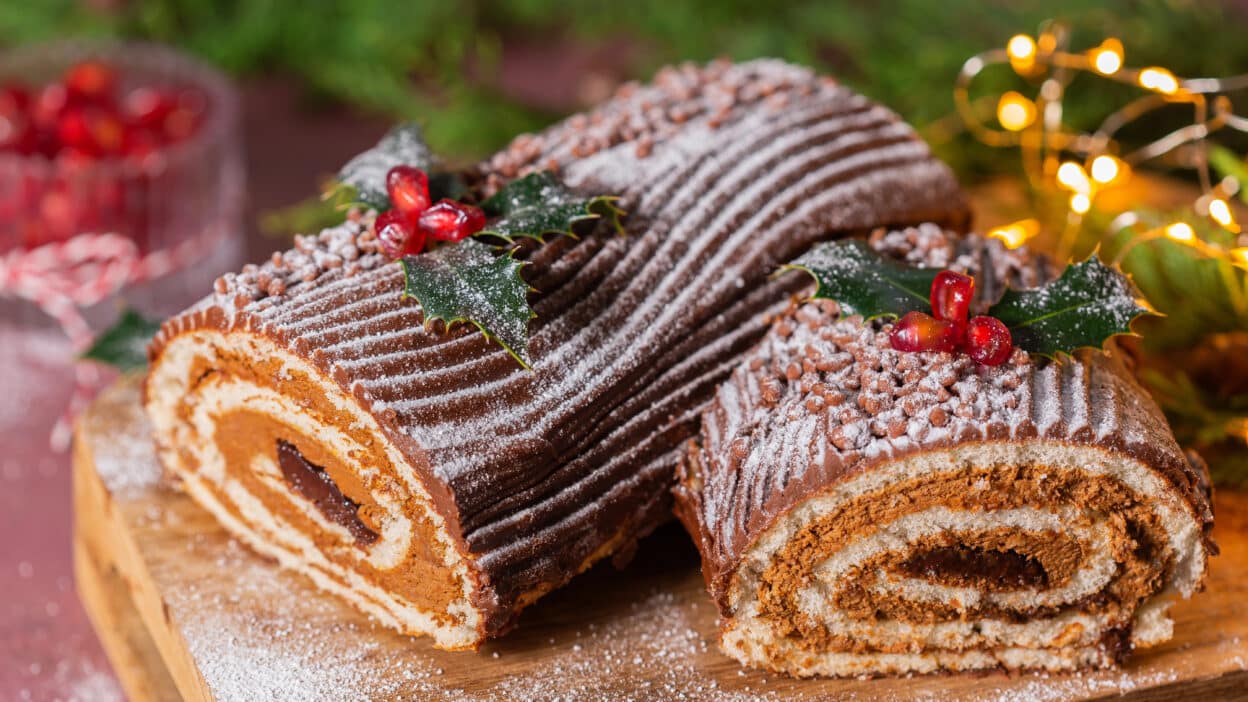 Festive Yule log cake with chocolate frosting, holly, and berries on a wooden board, dusted with powdered sugar.