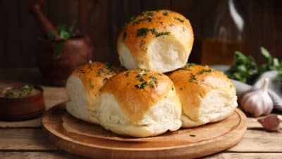 Freshly baked garlic herb rolls on a wooden platter, garnished with parsley, with rustic kitchen decor in the background.