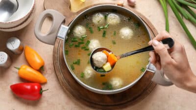 Hand serving meatball soup with veggies in a pot, surrounded by peppers, garlic, and herbs.