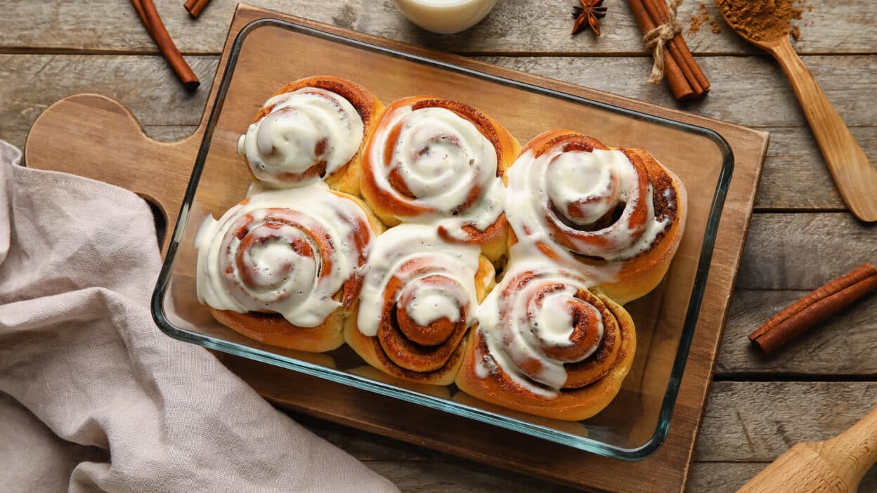 Freshly baked cinnamon rolls with creamy icing in a glass dish on a wooden board.