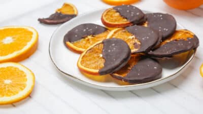 Chocolate-dipped dried orange slices on a white plate, garnished with sea salt, with fresh oranges nearby.
