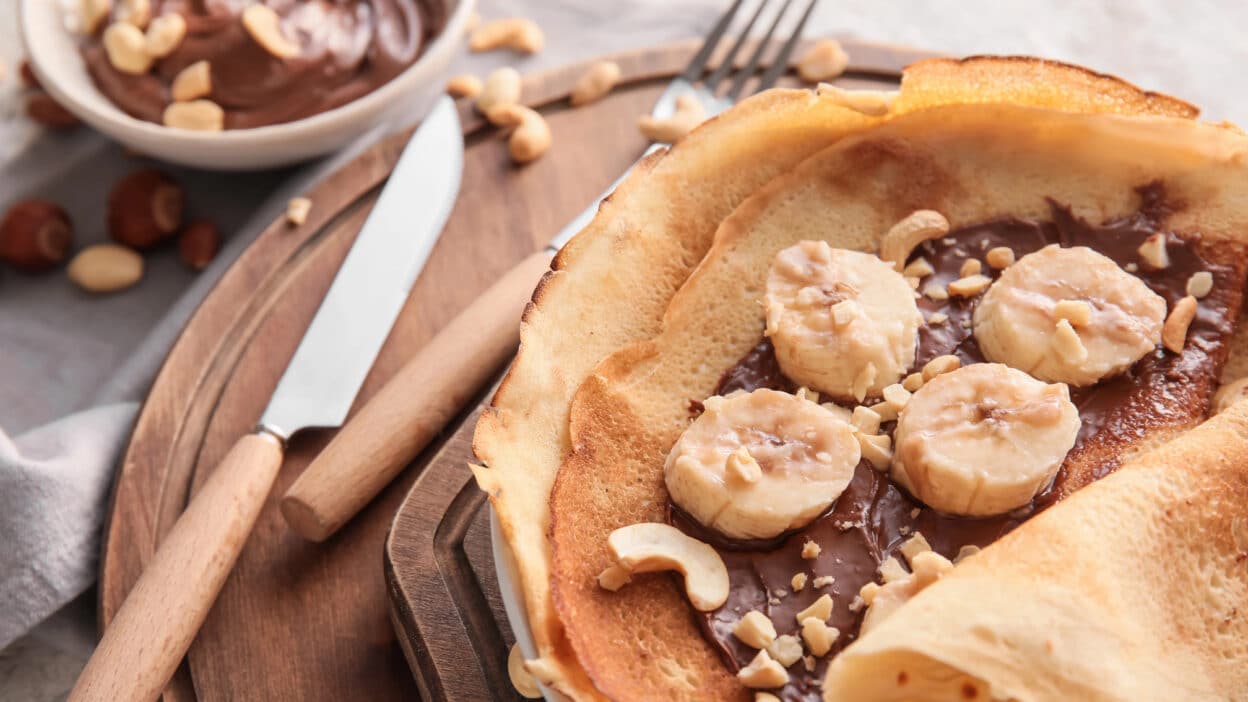 Crepes topped with banana slices, chocolate spread, and chopped nuts on a wooden board.