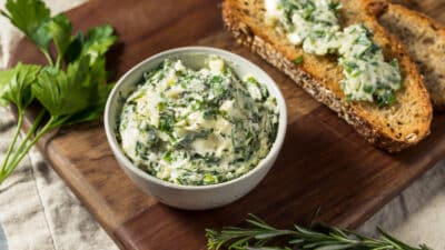 Creamy herb butter in a bowl with fresh bread on a wooden board, garnished with rosemary and parsley leaves.