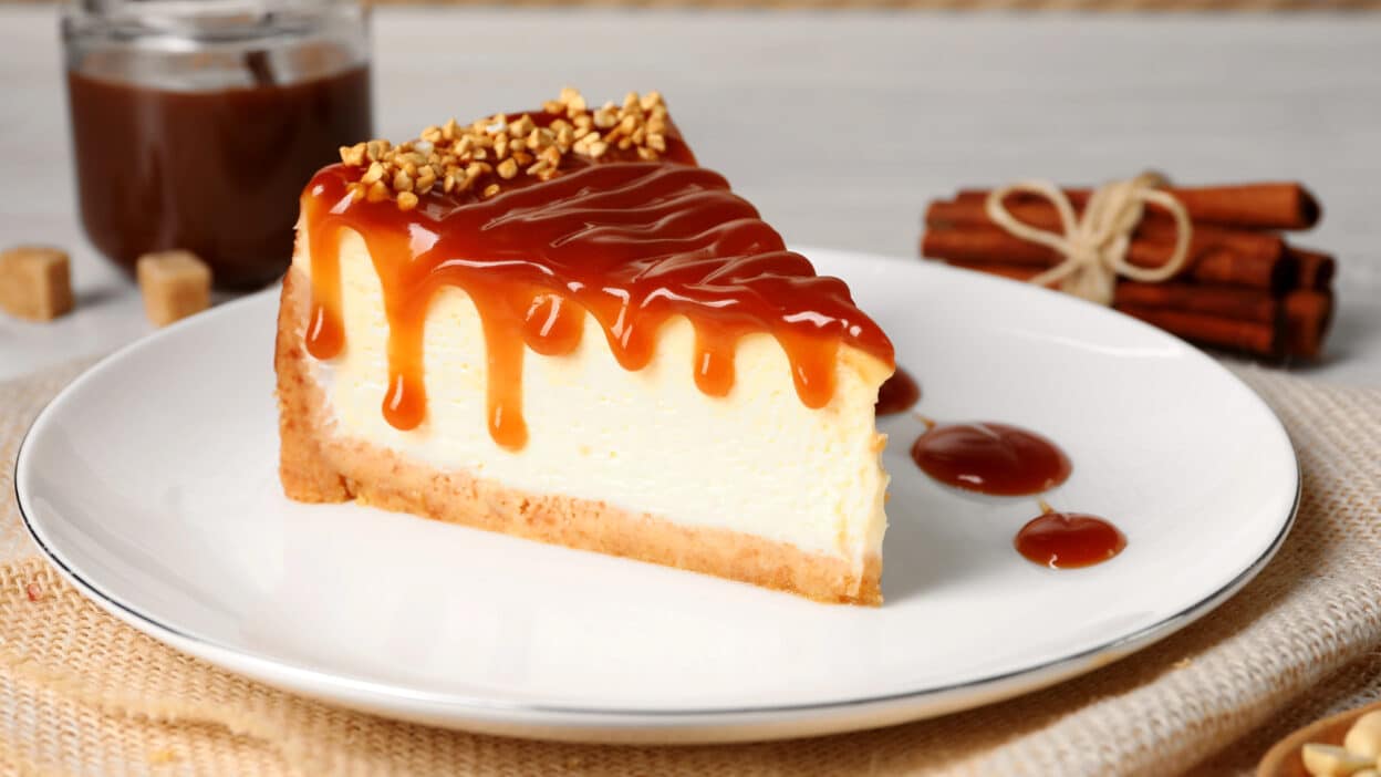 Slice of cheesecake with caramel sauce and nuts on top. Cinnamon sticks and caramel jar in the background.
