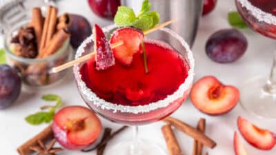 Red plum cocktail in a sugar-rimmed glass with fresh slices and mint garnish, surrounded by spices and ingredients.